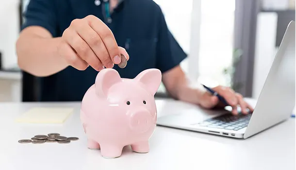 Ein Mensch wirft Geld in ein Sparschwein, während er vor einem Notebook sitzt.
